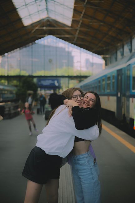 The image depicts two young women embracing each other in a joyful hug at Finchley Central station, with a train platform and a blue train in the background. The woman on the left is wearing glasses,
