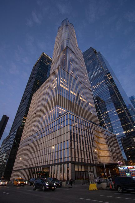 A tall modern skyscraper with a tiered, glass exterior illuminated in the evening, reflecting the surrounding cityscape. The building's reflective glass panels show the dusky sky and neighboring struc