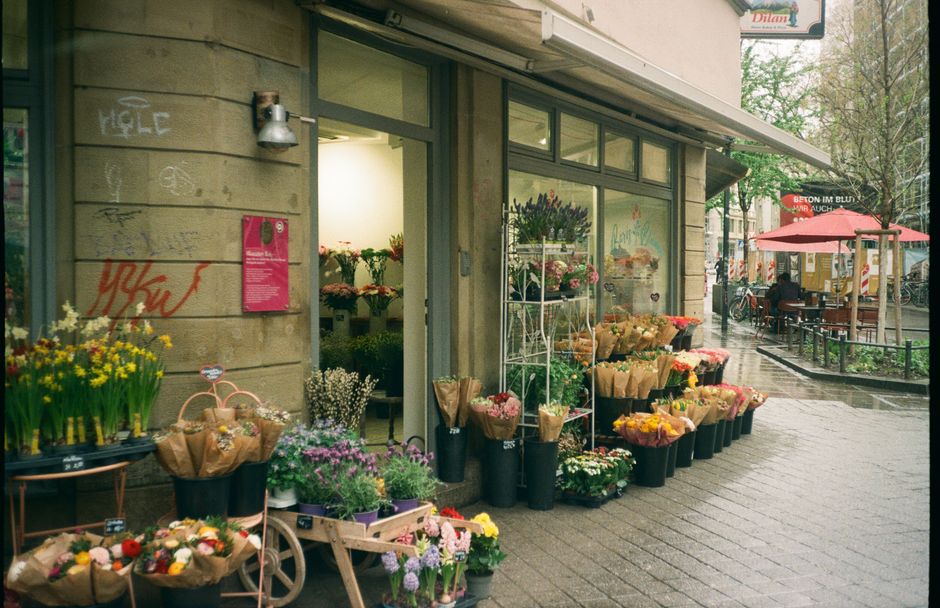 Exterior view of a flower shop along a wet pavement street, showcasing a variety of fresh floral bouquets and potted plants wrapped in brown paper and placed in black buckets, positioned outside the s