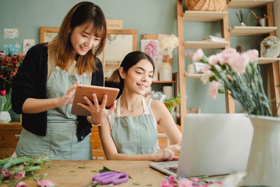 Two women in a flower shop with a light green interior, both wearing light green aprons. The woman on the left, standing, has shoulder-length brown hair with bangs and is smiling as she shows somethin