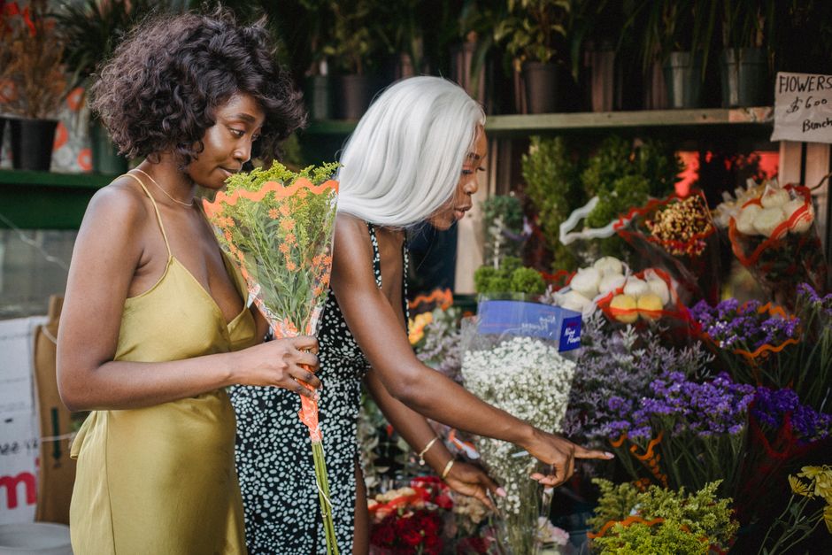 Two women are selecting fresh flowers at a flower shop in Finchley. The woman on the left, with dark curly hair, is holding a bouquet of orange and green flowers wrapped in clear cellophane, wearing a
