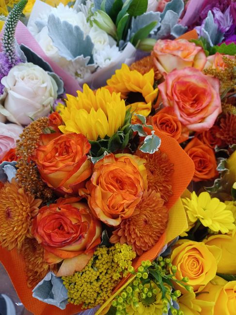 A vibrant floral arrangement featuring orange roses, yellow chrysanthemums, and pink spray roses, accented with green foliage and greyish-blue leaves. The flowers are tightly clustered and wrapped in 