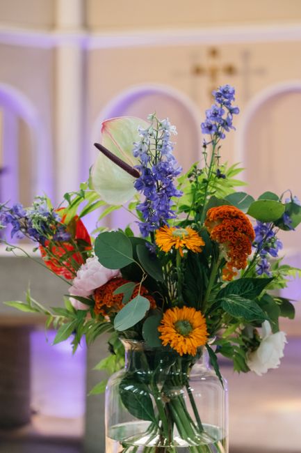 A vibrant floral arrangement in a clear glass vase featuring a variety of fresh flowers including purple delphiniums, orange calendulas, yellow daisies, and white carnations, accented with green folia
