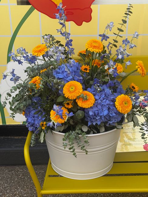 A vibrant floral arrangement featuring bright orange marigolds and yellow daisies mixed with clusters of blue hydrangeas and delicate blue delphiniums, all arranged in a white ceramic pot. The arrange